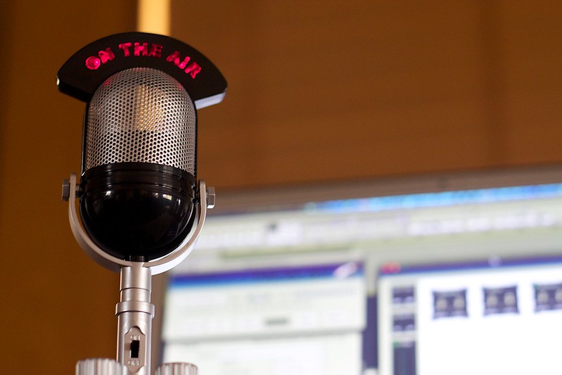 A radio microphone that says "on the air" in red sits in front of a blurred out computer in the background